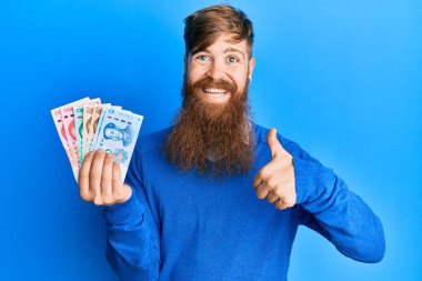 Genç İrlandalı kızıl adam Yuan 'ın Çin banknotlarını tutuyor mutlu ve pozitif gülümsüyor, başparmağıyla mükemmel ve onay işareti yapıyor. 