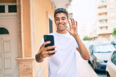 Genç Latin adam gülümseyerek şehirde akıllı telefon kullanarak video araması yapıyor..