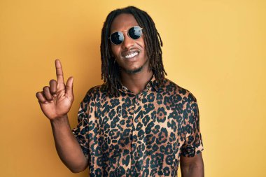 Saç örgülü, hayvan desenli tişörtlü ve güneş gözlüklü bir Afro-Amerikan adam bir fikir veya soruyla gülümsüyor ve mutlu bir yüzle işaret ediyor. 