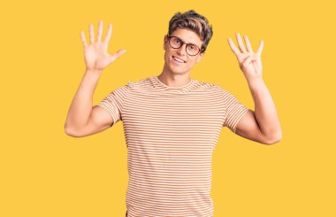 Genç, yakışıklı, günlük kıyafetler ve gözlük takan, dokuz numaralı parmaklarıyla işaret eden, kendinden emin ve mutlu bir şekilde gülümseyen bir adam.. 