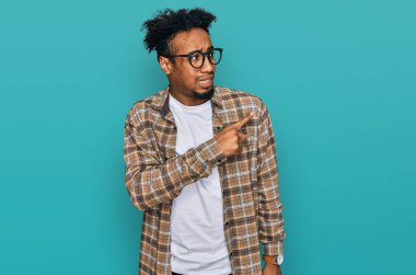 Young african american man with beard wearing casual clothes and glasses pointing aside worried and nervous with forefinger, concerned and surprised expression 