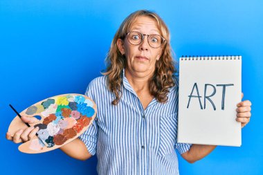 Orta yaşlı sarışın kadın elinde ressam paleti ve not defterinden habersiz ve kafası karışmış bir ifade tutuyor. şüphe kavramı. 
