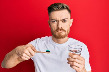 Genç kızıl saçlı adam diş fırçası ve bir bardak suyla depresif ve endişeli, sinirli ve korkmuş bir şekilde ağlıyordu. hüzünlü ifade. 