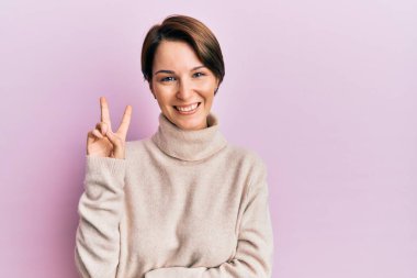 Kısa saçlı, kısa saçlı, kışlık süveter giyen, kameraya gülümseyen ve zafer işareti yapan genç esmer kadın. İki numara.. 