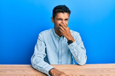Young handsome african american man wearing casual clothes sitting on the table smelling something stinky and disgusting, intolerable smell, holding breath with fingers on nose. bad smell 