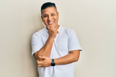 Gündelik beyaz tişört giyen genç Latin adam kameraya kendinden emin bakıyor kollarını kavuşturmuş ve çenesini kaldırmış bir şekilde gülümsüyor. Olumlu düşünmek. 