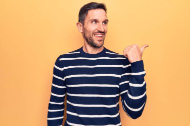 Sarı arka planda dikilen sıradan çizgili süveter giyen genç yakışıklı adam başparmağını yana kaldırıyor ve mutlu bir şekilde gülümsüyor.