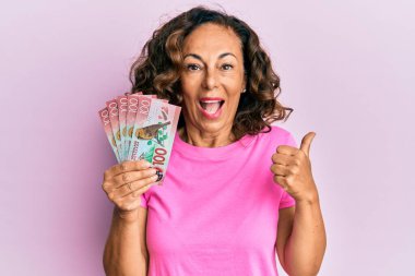 Orta yaşlı İspanyol bir kadın elinde 100 yeni Zelanda doları banknotuyla baş parmağıyla ağzını açıp gülümsüyor. 