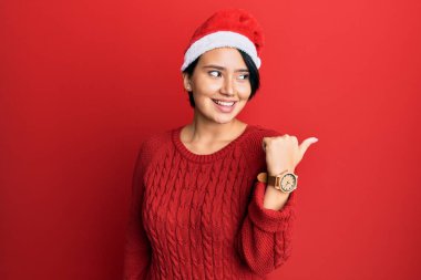 Kısa saçlı, güzel bir kadın Noel şapkası takıyor başparmağıyla yan tarafa bakıyor ve ağzı açık gülümsüyor. 