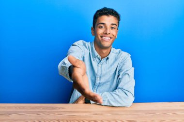 Young handsome african american man wearing casual clothes sitting on the table smiling cheerful offering palm hand giving assistance and acceptance. 