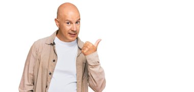 Orta yaşlı, kel, sıradan kıyafetler giyen, gülen yüzlü, gülümseyen ve baş parmağıyla yan tarafı gösteren bir adam.. 