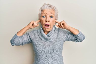 Yaşlı gri saçlı kadın parmaklarıyla kulaklarını kapatıyor. Şaşırmış ve şaşırmış bir ifade, korku ve heyecanlı bir yüz ifadesi var.. 