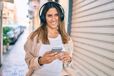 Beyaz kadın şehirde akıllı telefon ve kulaklık kullanarak mutlu bir şekilde gülümsüyor..