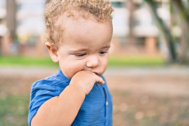 Üzgün küçük çocuk, güneşli bir günde parkta diş etlerine dokunduğu için parmaklarını dudaklarına koyuyor. Güzel sarı saçlı erkek bebek yeni bebek dişleri için acı çekiyor.