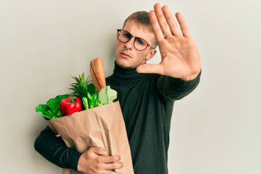 Elinde alışveriş poşeti olan beyaz bir genç adam dur işaretini ciddi ve kendinden emin bir ifadeyle yapıyor, savunma jesti. 