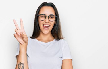 Gündelik beyaz tişört giyen, kameraya gülümseyen, gülümseyen ve parmaklarıyla zafer işareti yapan genç İspanyol kadın. İki numara.. 