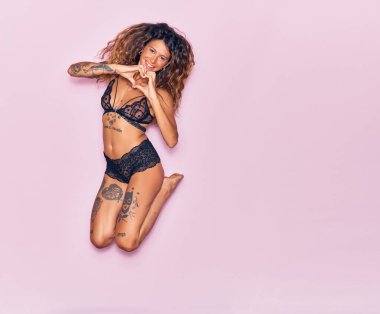 Dövmeli, iç çamaşırlı, mutlu gülümseyen genç bir kadın. Yüzünde tebessümle zıplıyor, kalp şeklinde, elleri izole pembe arka planda.