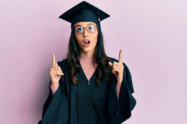 身穿毕业典礼帽和礼服的年轻女子惊奇而惊奇地抬起头 用手指和胳膊指指着 — 图库照片