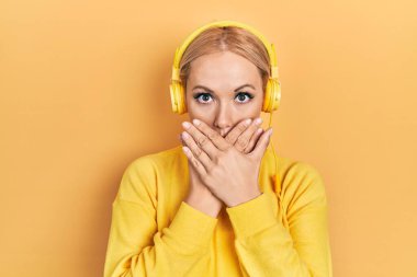 Kulaklık kullanarak müzik dinleyen genç sarışın kadın yanlışlıkla elleriyle ağzını kapatırken şok oldu. gizli kavram. 