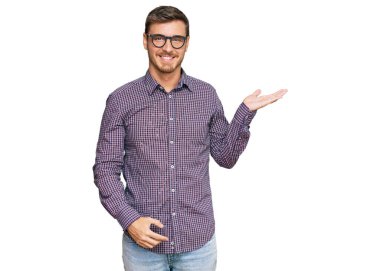 Yakışıklı beyaz bir adam günlük kıyafetler ve gözlük takıyor neşeli bir şekilde gülümsüyor ve avuç içi kameraya bakıyor.. 