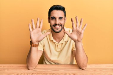 Latin asıllı genç adam, gündelik giysiler içinde masada oturuyor ve 10 numaralı parmaklarıyla onu gösterirken kendinden emin ve mutlu bir şekilde gülümsüyor.. 
