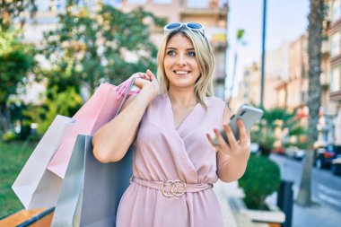 Genç sarışın kadın gülümsüyor alışveriş torbaları tutuyor ve şehirde akıllı telefon kullanıyor..