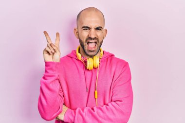 Young bald man wearing gym clothes and using headphones smiling with happy face winking at the camera doing victory sign. number two. 