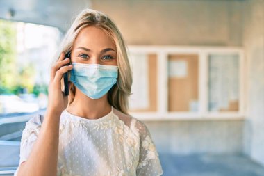Tıbbi maske takan genç beyaz kız şehrin caddesinde akıllı telefondan konuşuyor..