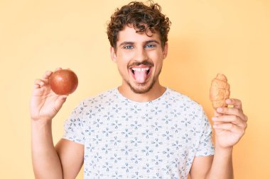 Kıvırcık saçlı, elma ve kruvasan tutan genç beyaz adam komik bir ifadeyle dilini dışarı çıkartıyor.. 