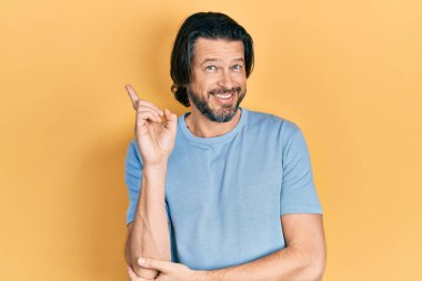 Orta yaşlı, günlük kıyafetler giyen, yüzünde kocaman bir gülümseme olan beyaz bir adam, kameraya el ve parmak ile bakıyor.. 