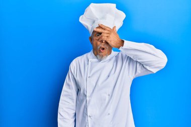 Orta yaşlı, gri saçlı, profesyonel aşçı üniforması giyen, şapkası olan, yüzünü ve gözlerini kapayarak utangaç ifadesiyle parmaklarından bakan bir adam.. 
