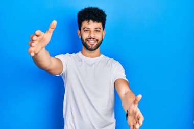 Sakallı, beyaz tişörtlü genç bir Arap kameraya sarılıp gülümsüyor. Mutluluğu kucaklayan neşeli bir ifade.. 