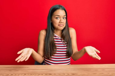 Gündelik giysiler giymiş güzel İspanyol bir kadın masanın üzerinde elleri ve kolları havada, kafası karışmış bir şekilde oturuyor. şüphe kavramı. 