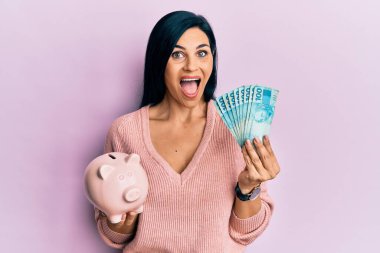 Young caucasian woman holding brazil real banknotes and piggy bank celebrating crazy and amazed for success with open eyes screaming excited. 