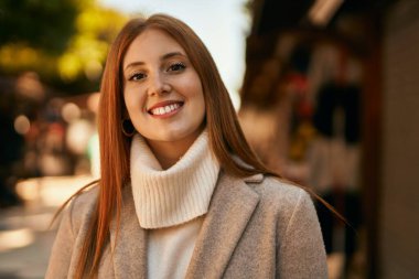 Genç kızıl kız gülümsüyor, mutlu bir şekilde şehirde duruyor..