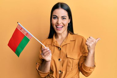 Young brunette woman holding belarus flag pointing thumb up to the side smiling happy with open mouth 