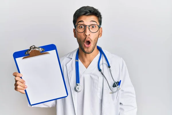 Jovem Homem Bonito Vestindo Médico Estetoscópio Segurando Prancheta Assustado Surpreso — Fotografia de Stock