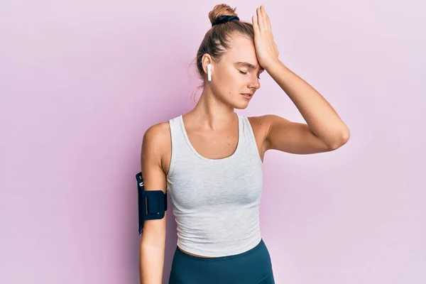 Beautiful blonde sport woman wearing arm band and earphones surprised with hand on head for mistake, remember error. forgot, bad memory concept.