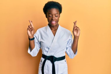 Young african american girl wearing karate kimono and black belt gesturing finger crossed smiling with hope and eyes closed. luck and superstitious concept. 