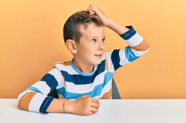 Sevimli beyaz çocuk, gündelik giysiler içinde masaya oturmuş gülümsüyor, el hareketleriyle saçlarına dokunuyor, çekici ve modaya uygun poz veriyor. 