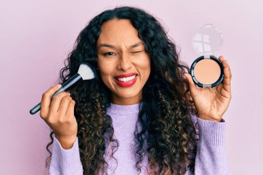 Makyaj fırçası tutan genç Latin kadın kameraya seksi bir ifadeyle bakıyor, neşeli ve mutlu bir yüzle.. 