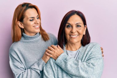 İspanyol anne ve kızı mutlu bir şekilde gülümsüyor ve soyutlanmış pembe arka planda duruyorlar..