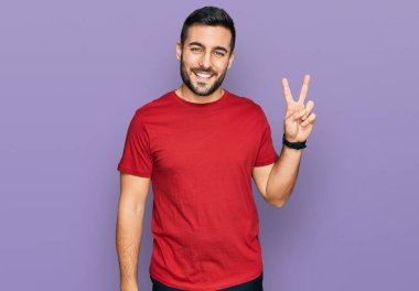 Gündelik kıyafetler giyen, gülümseyen ve kameraya göz kırpan genç İspanyol adam zafer işareti yapıyor. İki numara.. 