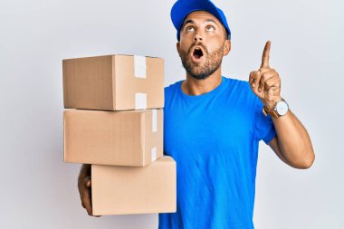 Sakallı, kurye üniformalı yakışıklı bir adam teslimat paketlerini tutarken şaşkın ve şaşırmış bir şekilde yukarı bakıyor ve parmaklarıyla işaret ediyor ve kollarını kaldırıyor.. 