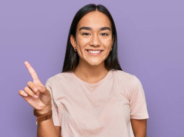 Genç Asyalı kadın gündelik giysiler giyip bir numarayı gösterip işaret ederken kendine güvenen ve mutlu bir şekilde gülümsüyor.. 