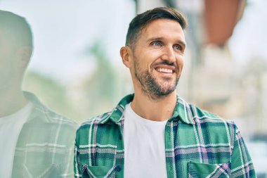 Genç beyaz adam gülümseyerek şehirde mutlu mesut duruyor..