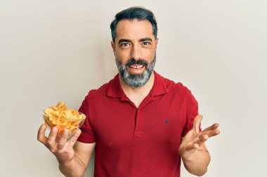 Orta yaşlı, sakallı, elinde patates cipsi tutan mutlu bir gülümsemeyle başarıyı kutlayan ve elini kaldıran bir adam. 