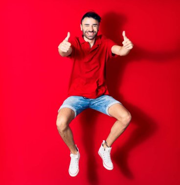 Şapkalı genç teslimatçı mutlu gülümsüyor. Yüzünde tebessümle zıplıyor. Tamam işareti yapıyor. Kırmızı arka plan üzerinde başparmaklar yukarı doğru.