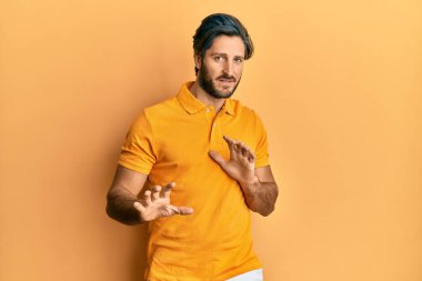 Latin asıllı genç bir adam günlük sarı tişört giyiyor tiksindirici bir ifade takınıyor ve tiksindirici bir yüz ifadesi takınıyor. Eller havada. 