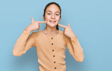 Güzel esmer, sıradan bir süveter giyen neşeli bir şekilde gülümseyen ve parmaklarıyla ve ağzıyla işaret eden küçük bir kız. diş sağlığı kavramı. 